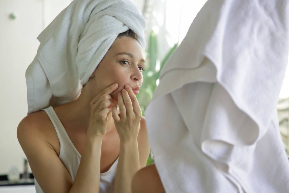 A woman in a towel head wrap looks in the mirror at her acne